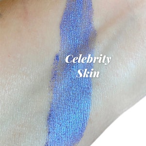 May include: A close-up of a blue iridescent eyeshadow swatch on skin. The text "Celebrity Skin" is written in white on the swatch.
