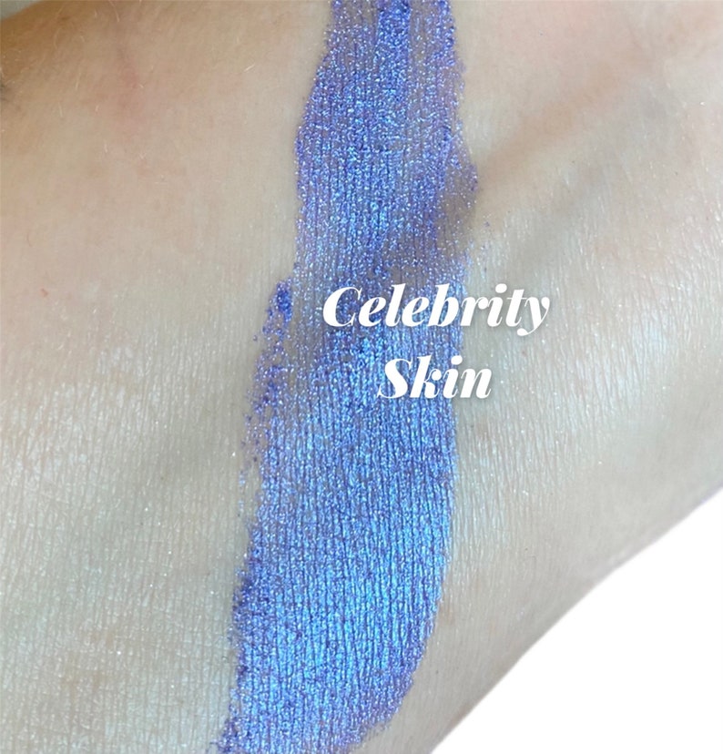 May include: A close-up of a blue iridescent eyeshadow swatch on skin. The text "Celebrity Skin" is written in white on the swatch.