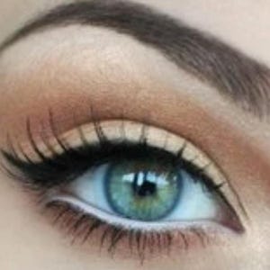 May include: Close-up of an eye with teal iris, dark lashes, and a defined brow. The eye makeup includes a black eyeliner and a light brown eyeshadow. The skin around the eye is a light peach color.