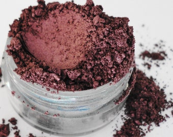 May include: A close-up of a small jar filled with a deep burgundy eyeshadow powder. The powder has a slightly shimmery finish and is spilling out of the jar.