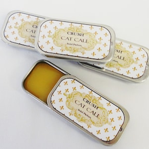 Op de afbeelding: Vier rechthoekige metalen blikken met vaste parfum, waarvan één open, met een lichtgele vaste stof erin. De deksels zijn wit met een gouden fleur-de-lis patroon en de tekst "CRUSH CAT CALL Solid Parfum".