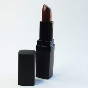 May include: A dark burgundy lipstick in a square black casing with the cap removed and placed beside it. The lipstick bullet is extended, showing a deep, rich red-brown color with a smooth, slightly glossy finish on the tip of the product.