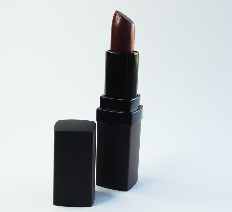 May include: A dark burgundy lipstick in a square black casing with the cap removed and placed beside it. The lipstick bullet is extended, showing a deep, rich red-brown color with a smooth, slightly glossy finish on the tip of the product.