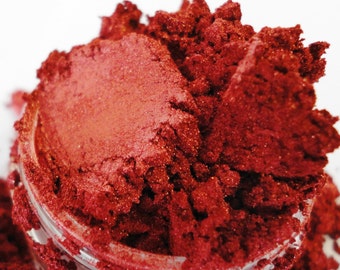May include: Close-up of a red mica powder in a small glass jar. The powder is fine and shimmery, with a slightly metallic sheen.