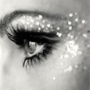 May include: Close-up, black and white image of an eye with long eyelashes. Silver glitter is scattered around the eye, creating a sparkling effect. The eye is open, and the iris is visible.