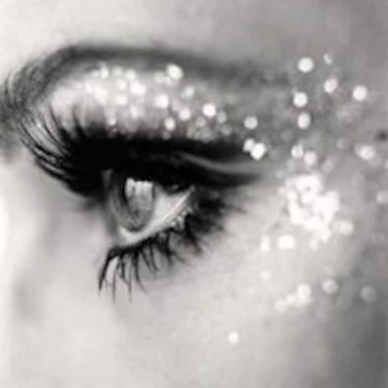 May include: Close-up, black and white image of an eye with long eyelashes. Silver glitter is scattered around the eye, creating a sparkling effect. The eye is open, and the iris is visible.