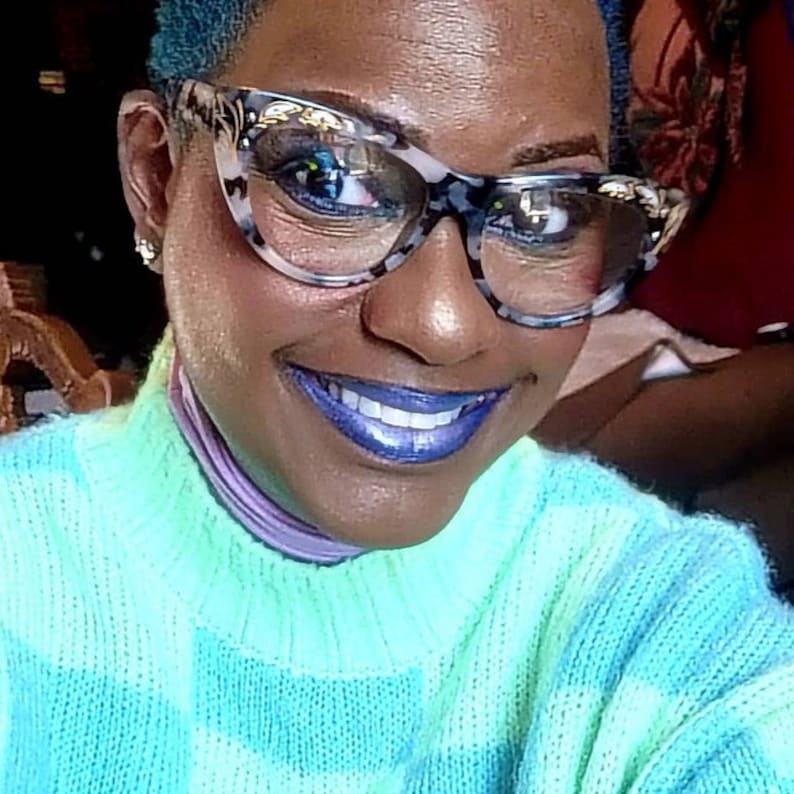 May include: A woman wearing a blue and green patterned sweater. She is wearing glasses with a gold trim and has blue lipstick.