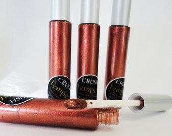 May include: Four tubes of shimmering copper-colored lip gloss with silver caps. One tube is open, revealing the applicator and the gloss. The label reads "CRUSH Tempesta". The gloss has a metallic finish.