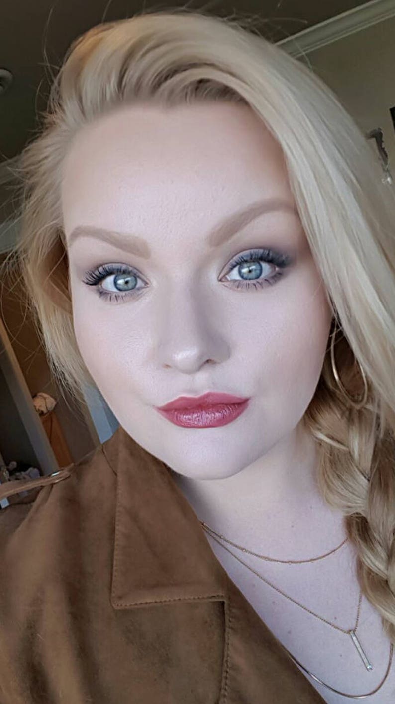Puede incluir: Una persona con cabello rubio peinado en una trenza lateral, vistiendo una chaqueta de ante marr&oacute;n con cuello, pendientes de aro dorados y un collar dorado de varias capas. El maquillaje incluye cejas definidas, sombra de ojos azul y labial rosa.