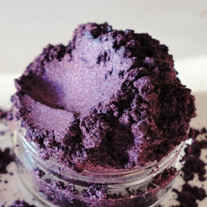 May include: A close-up of a small jar filled with a deep purple iridescent mica powder. The powder has a shimmery, almost holographic finish.