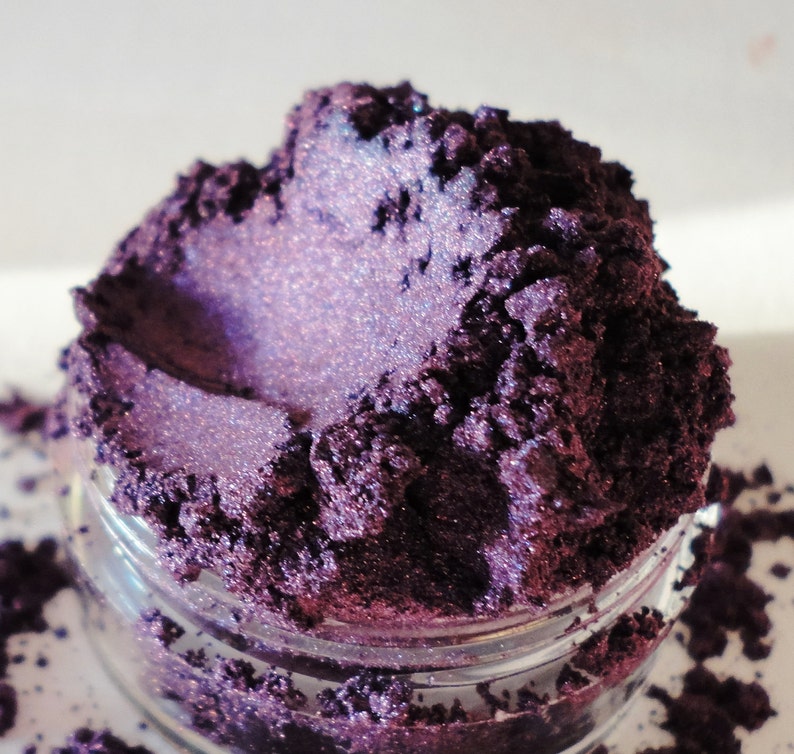 May include: A close-up of a small jar filled with a deep purple iridescent mica powder. The powder has a shimmery, almost holographic finish.