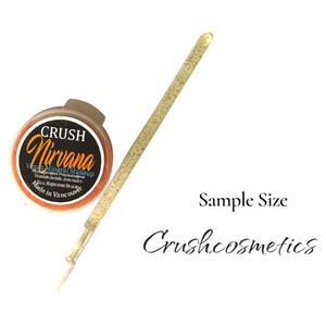 May include: A small round container of Crush Nirvana vegan mineral makeup with a black label and orange text, accompanied by a long, thin, gold glitter-covered applicator brush. The text Sample Size and Crushcosmetics appear in black.