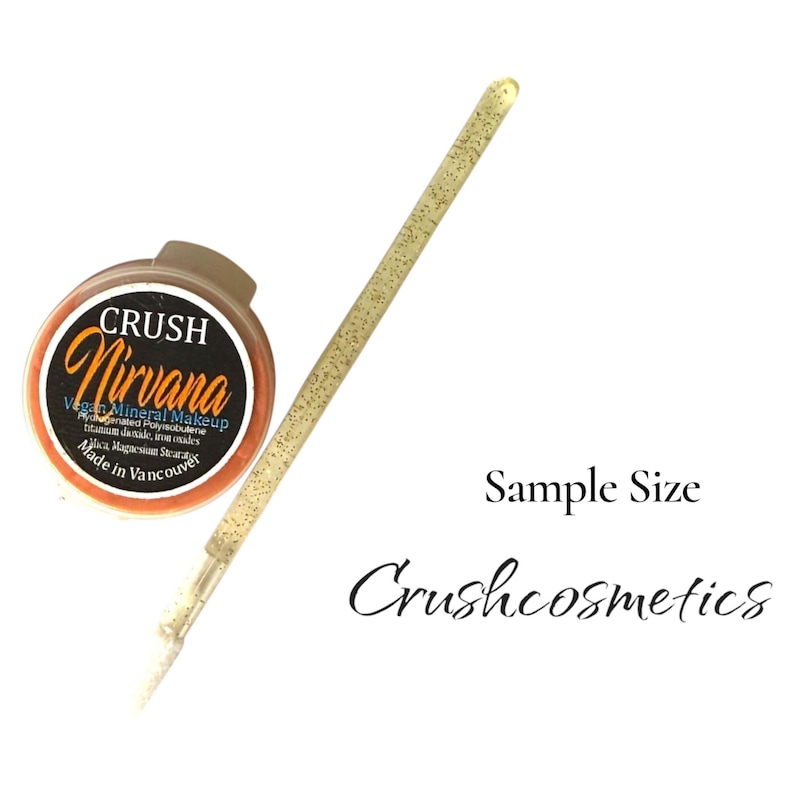 May include: A small round container of Crush Nirvana vegan mineral makeup with a black label and orange text, accompanied by a long, thin, gold glitter-covered applicator brush. The text Sample Size and Crushcosmetics appear in black.