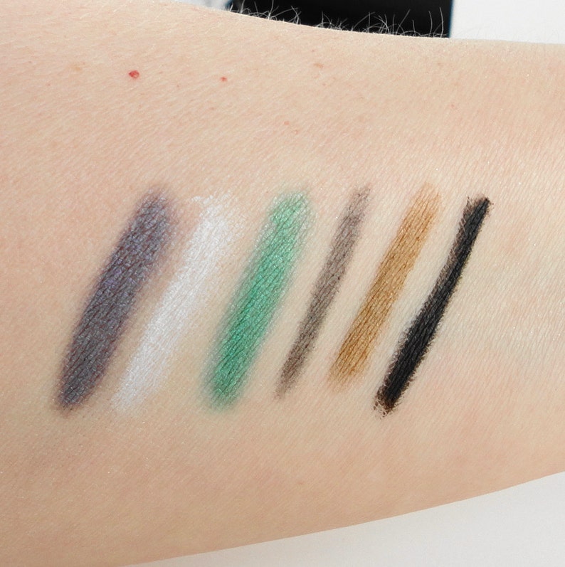 May include: A close-up of seven eyeliner pencil swatches on a person's arm. The colours are grey, white, green, grey, brown, black, and black.