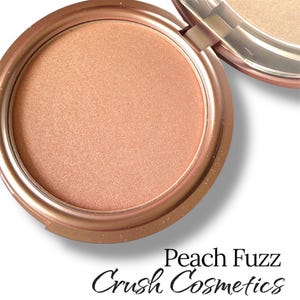 May include: A round compact of pressed powder in a shimmering peach tone, housed in a metallic bronze case with a hinged lid. The text Peach Fuzz and Crush Cosmetics is printed in black below the open container on a plain white background.