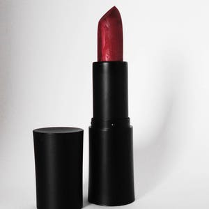 Puede incluir: Un labial con una barra de color rojo intenso y textura ligera extendida desde un estuche cil&iacute;ndrico negro mate. La tapa negra a juego est&aacute; colocada en posici&oacute;n vertical a la izquierda del tubo principal, ambos con un acabado negro liso.