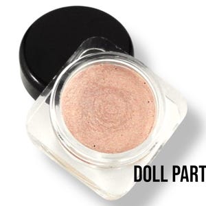 Könnte beinhalten: Ein kleines, klares Glas mit schwarzem Deckel, das einen schimmernden, hellrosa Creme-Lidschatten enthält. Das Glas ist mit "DOLL PARTS" beschriftet.