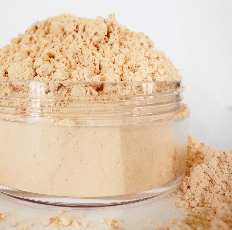 May include: A clear plastic jar filled with loose light beige powder. The powder is spilling out of the jar onto a white surface.