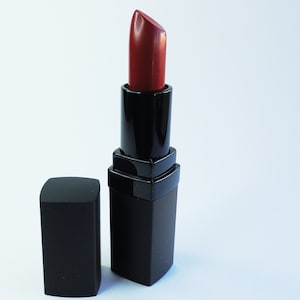 May include: A tube of lipstick with a deep red pigment extended from its black rectangular casing. The matching black cap is positioned upright next to the base of the lipstick tube. The product features a smooth, matte finish on the exterior.