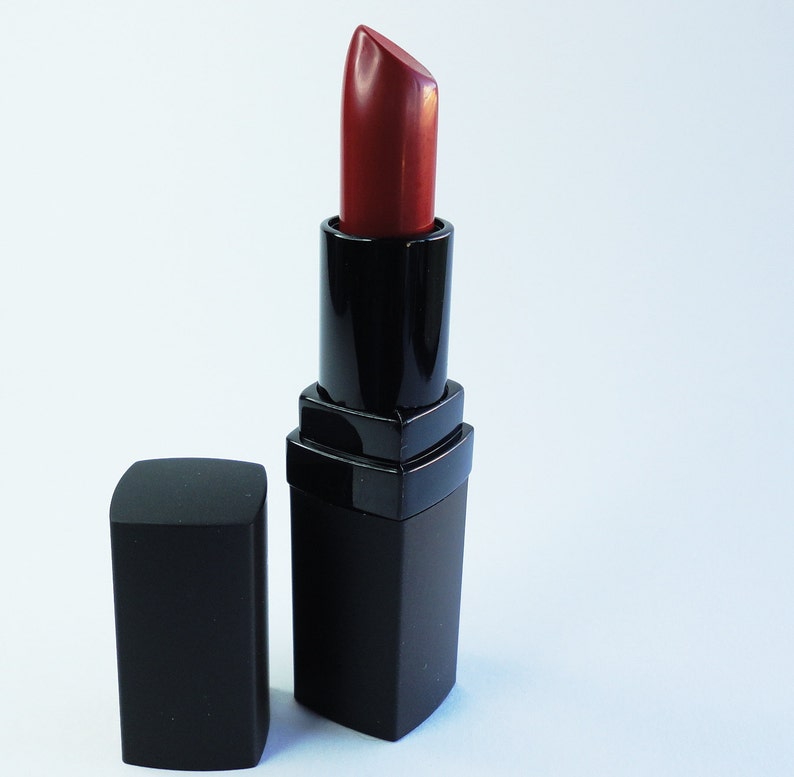 May include: A tube of lipstick with a deep red pigment extended from its black rectangular casing. The matching black cap is positioned upright next to the base of the lipstick tube. The product features a smooth, matte finish on the exterior.