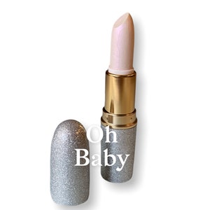 Puede incluir: Un tubo de lápiz labial de brillo plateado con una tapa dorada y una bala de lápiz labial blanco iridiscente. El tubo tiene el texto "Oh Baby" impreso.