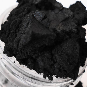 May include: A close-up of a small jar filled with black powder. The powder is fine and has a slightly grainy texture.