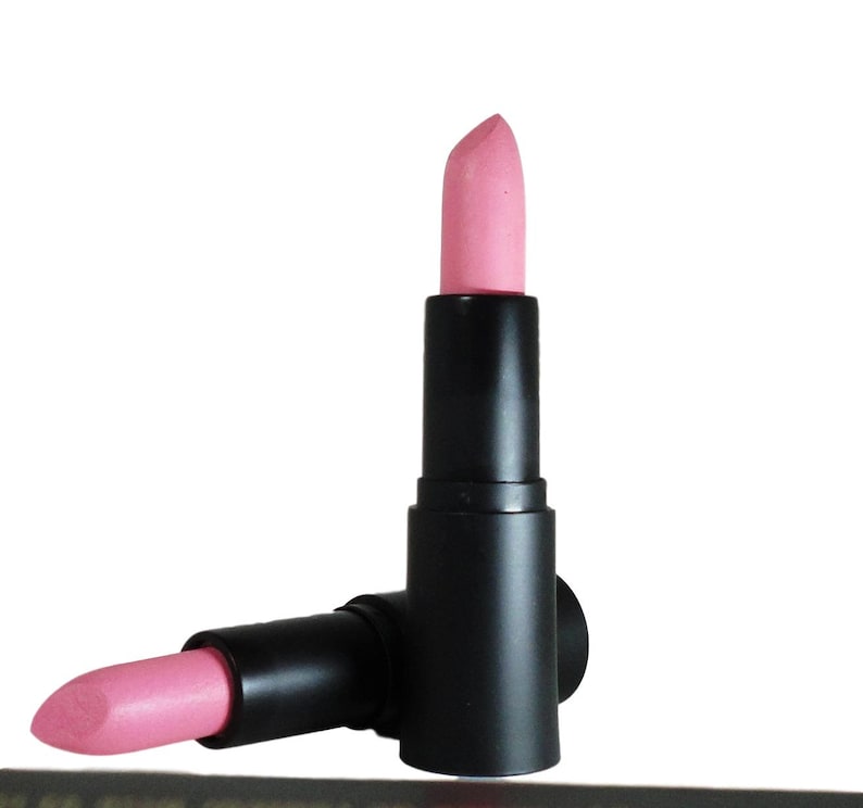 May include: Two lipstick tubes with matte black cylindrical casings. One tube stands vertically with the light pink lipstick bullet extended, while the second tube lies horizontally at the base with its light pink lipstick bullet partially exposed.