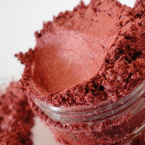 May include: A close-up of a small jar filled with a deep red, shimmery loose pigment powder. The powder is spilling out of the jar, creating a textured, uneven surface.