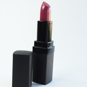 May include: A tube of lipstick with a mauve-toned, satin-finish bullet extended from a black, rectangular casing. The matching black cap is positioned upright next to the base of the lipstick tube, which features a smooth, matte-textured exterior.