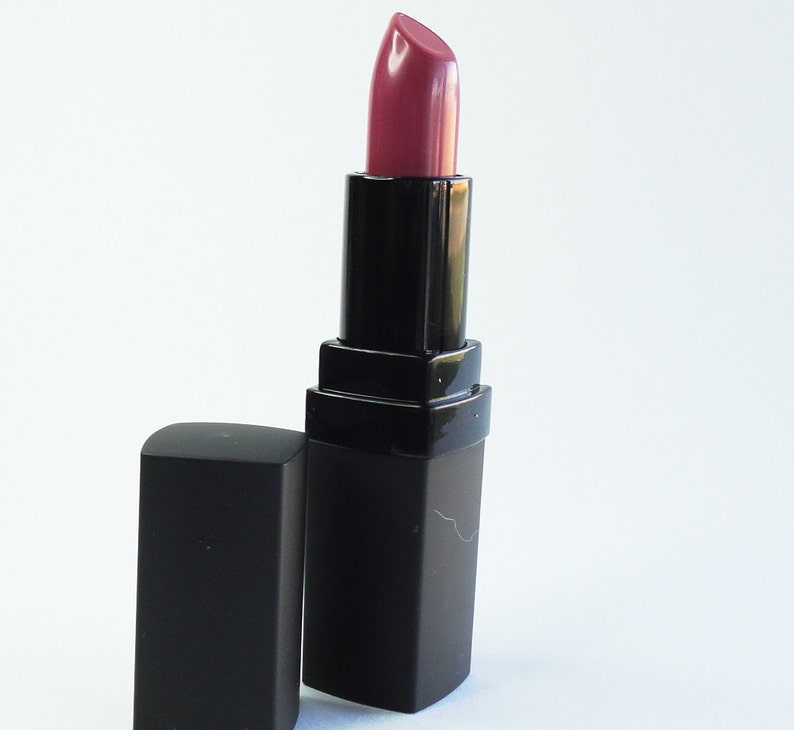 May include: A tube of lipstick with a mauve-toned, satin-finish bullet extended from a black, rectangular casing. The matching black cap is positioned upright next to the base of the lipstick tube, which features a smooth, matte-textured exterior.