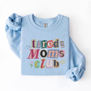 May include: A light blue sweatshirt with the words "tired Moms Club" in a colorful, cut-out style design. The design includes a coffee cup, a flower, a cloud, and a star. The sweatshirt is made of a soft material.
