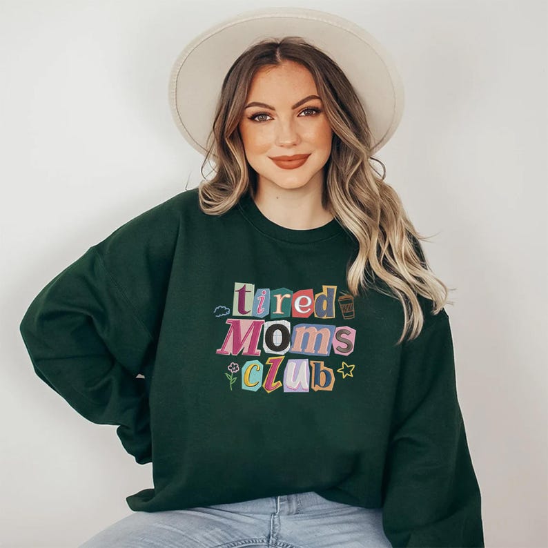 May include: Dark green sweatshirt with the text "Tired Moms Club" in colorful, blocky letters. The sweatshirt has a crew neck and long sleeves. A woman is wearing the sweatshirt.