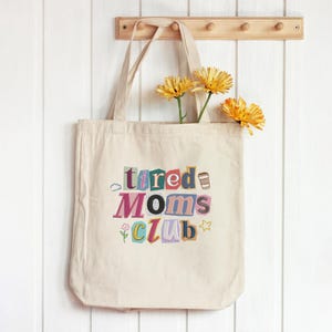 May include: A beige canvas tote bag with the words "tired Moms Club" in colorful, cut-out style letters. The bag has a simple design and is hanging on a wooden rack. Yellow flowers are placed inside the bag.