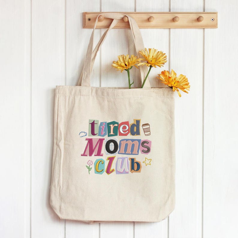 May include: A beige canvas tote bag with the words "tired Moms Club" in colorful, cut-out style letters. The bag has a simple design and is hanging on a wooden rack. Yellow flowers are placed inside the bag.