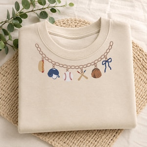 May include: A beige crewneck t-shirt featuring an embroidered graphic of a gold chain necklace. The chain includes charms shaped like a hot dog, a blue baseball helmet, a white baseball, two crossed wooden bats, a brown baseball glove, and a blue bow.