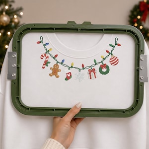 May include: A white sweatshirt secured in a green embroidery hoop featuring a festive embroidered garland design. The garland consists of colorful Christmas lights, a candy cane, a gingerbread man, a stocking, a snowflake, a gift, and a wreath.