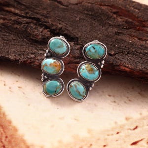 May include: A pair of silver earrings, each featuring three oval-shaped turquoise stones with brown matrix patterns. The stones are set in polished silver bezels with small silver bead accents between each stone, creating a vertical, curved design.