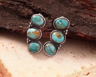 May include: A pair of silver earrings, each featuring three oval-shaped turquoise stones with brown matrix patterns. The stones are set in polished silver bezels with small silver bead accents between each stone, creating a vertical, curved design.