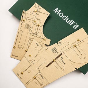 Puede incluir: Plantillas de costura de madera clara con cierres metálicos dorados y marcas grabadas. Las piezas tienen etiquetas como F1, F4 Shoulder, F5 Arm Hole y Bk Crotch. El conjunto se muestra sobre una caja de color verde oscuro con la etiqueta ModulFit.