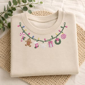 May include: A beige crewneck sweatshirt featuring an embroidered garland of green wire with colorful Christmas lights and hanging ornaments including a candy cane, gingerbread man, stocking, snowflake, gift box, wreath, and striped ball.