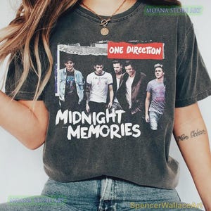 Pode incluir: Uma t-shirt de manga curta em cinzento-carvão com uma impressão gráfica da banda One Direction. O design inclui o texto One Direction e Midnight Memories, juntamente com uma imagem fotográfica dos cinco membros da banda em roupa casual.