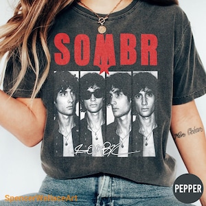May include: A charcoal gray short-sleeve t-shirt featuring a red distressed text graphic that reads SOMBR at the top. Below the text are four black and white portrait panels of the same person, with the word SOMBR written in white script below.