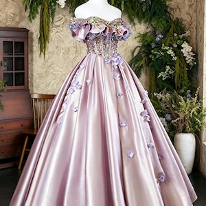 May include: A floor-length off-the-shoulder ball gown in a shimmering mauve satin fabric. The bodice features intricate beadwork and sequins, while the neckline is adorned with ruffled fabric and small purple floral appliqués scattered throughout.