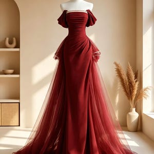 May include: A floor-length, deep red gown featuring an off-the-shoulder neckline with short puff sleeves, a fitted bodice with subtle ruching, and a sheer tulle overlay that extends into a long train, displayed on a white dress form mannequin.