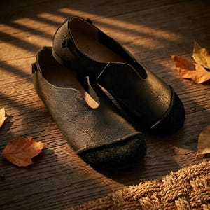 May include: A pair of black leather flats with a rounded toe and a small heel. The shoes have a t-strap design and are set on a wooden surface with autumn leaves.