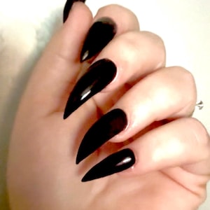 Könnte beinhalten: Nahaufnahme einer Hand mit langen, glänzenden schwarzen Stiletto-Nägeln. Die Nägel sind spitz und reflektieren das Licht, was einen dramatischen Look erzeugt. Die Maniküre ist einfarbig schwarz.