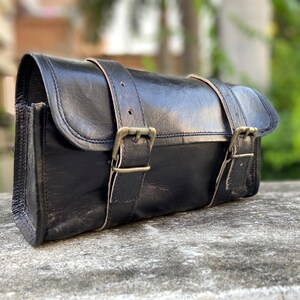 Peut inclure: Sacoche en cuir noir avec deux sangles bouclées sur le dessus. Le sac est de forme rectangulaire avec des bords arrondis et une fermeture à rabat. La quincaillerie est de couleur laiton et le cuir semble vieilli.