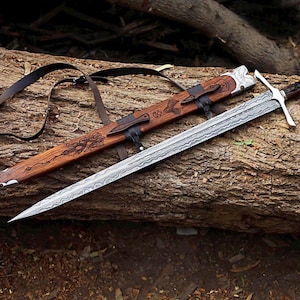 May include: A Damascus steel sword with a silver crossguard and a dark wood handle featuring silver accents. The matching wooden scabbard is engraved with intricate patterns and includes a black leather strap for carrying the weapon.