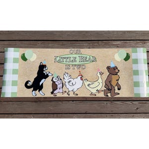 May include: A banner with the text "OUR LITTLE BEAR IS TWO" and illustrations of a cat, owl, chicken, duck, and bear, all wearing party hats. Green and white balloons are on either side of the text. The banner has a light brown background with green and white checkered edges.