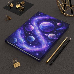 Op de afbeelding: Een hardcover notitieboek met een ruimtethema. De omslag toont een melkwegstelsel met planeten in blauw, paars en wit. Gouden en zwarte potloden en paperclips zijn ook zichtbaar.